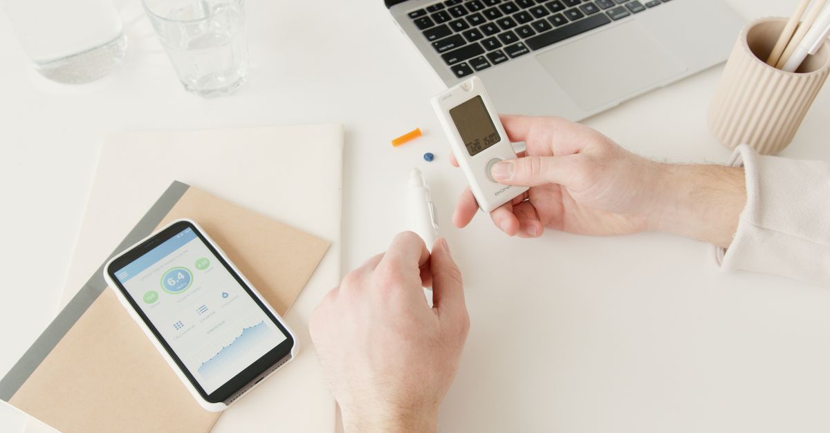 Person checking blood sugar with glucometer on a desk with smartphone and laptop nearby.