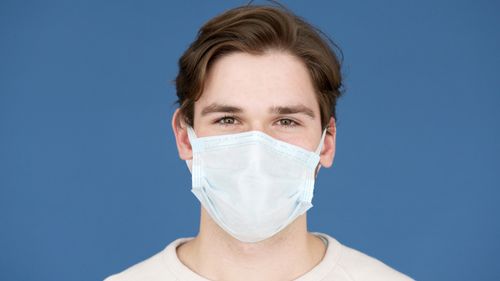 Free Portrait of a young man wearing a face mask against a blue background, emphasizing pandemic safety. Stock Photo