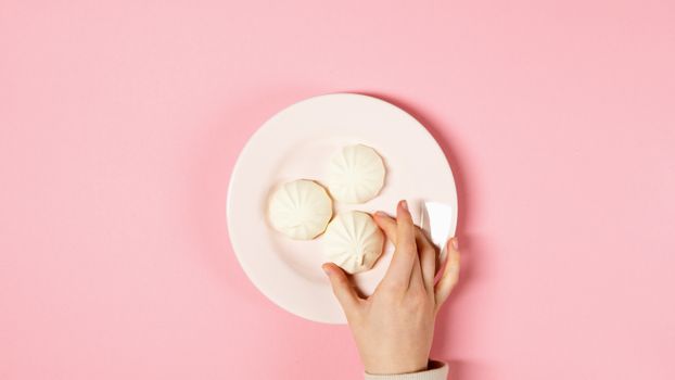 A hand holds a ceramic plate of white meringues against a light pink background.