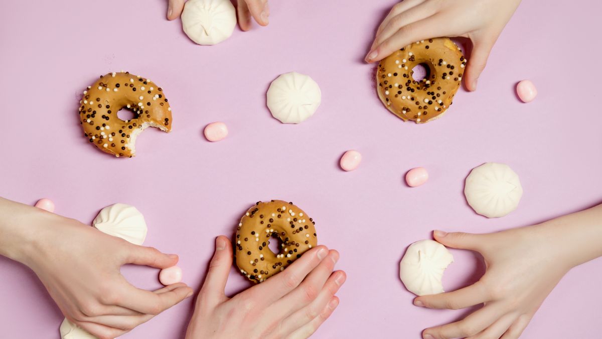 Top view of hands holding donuts with sprinkles and candies on pastel background.