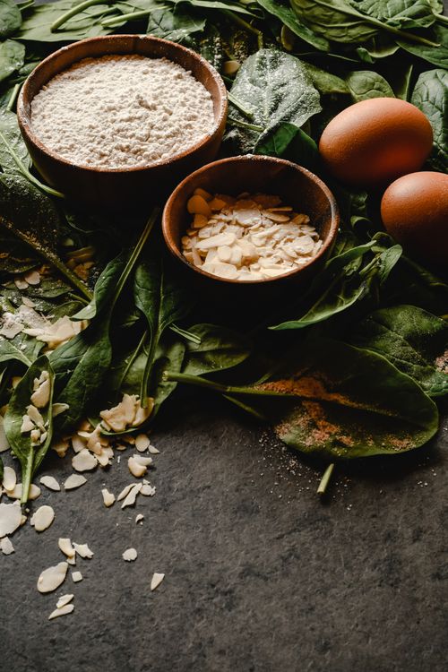 Free Close-up of wooden bowls with flour and almonds, surrounded by fresh spinach and eggs on a dark surface. Stock Photo