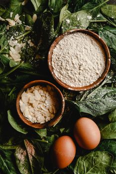Top view of fresh spinach, eggs, flour, and almond flakes set for preparing spinach pancakes.