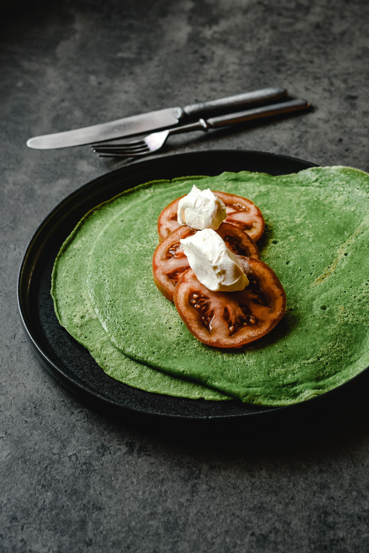 Bright green spinach crepes on a black plate topped with tomato slices and cream cheese