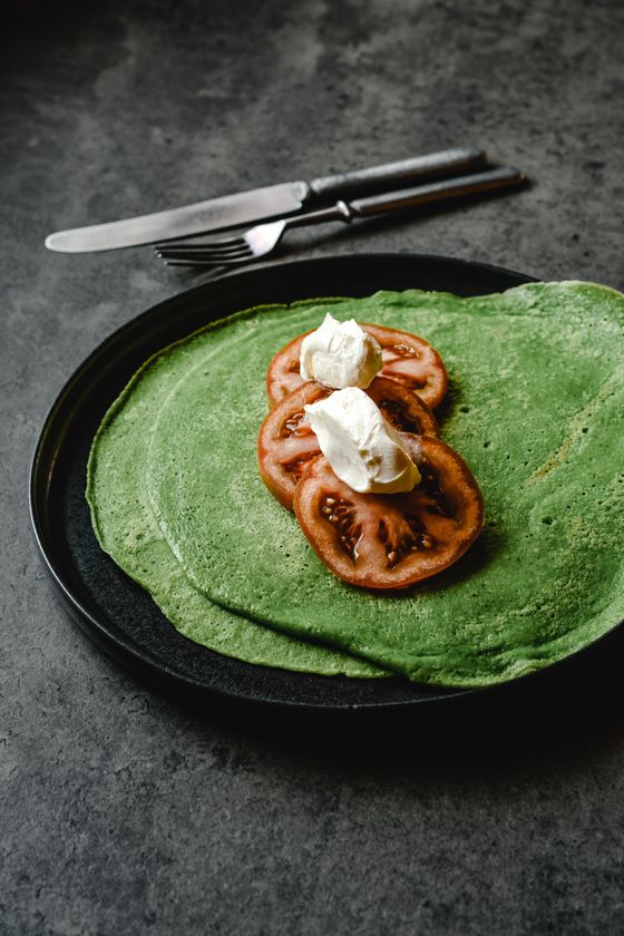 Bright green spinach crepes on a black plate topped with tomato slices and cream cheese