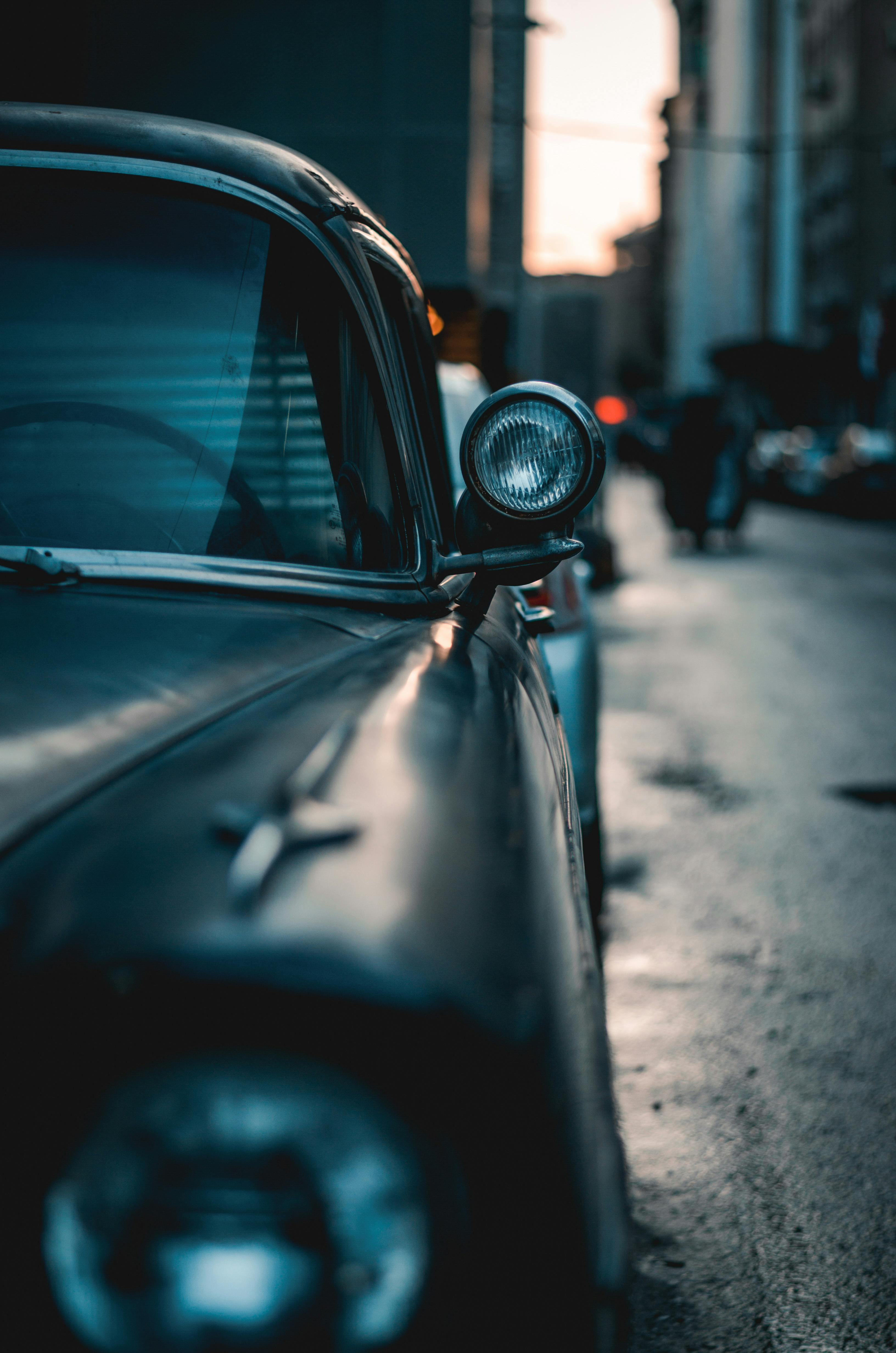 Close-up of a vintage car bumper and headlight on a rainy city street during twilight.