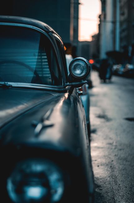 Close-up of a vintage car bumper and headlight on a rainy city street during twilight.