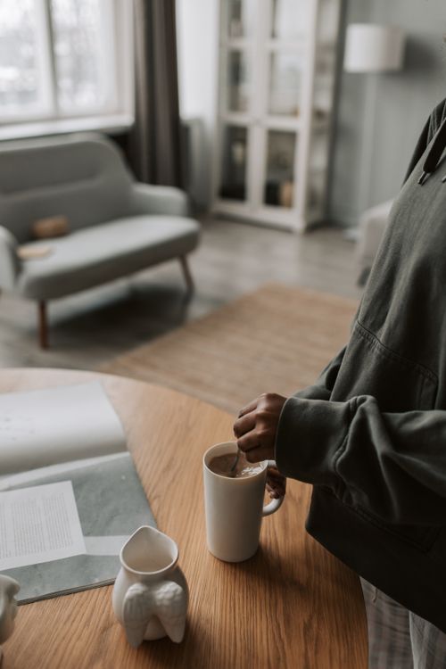 Free A person stirs coffee in a mug on a wooden table inside a modern living room. Stock Photo