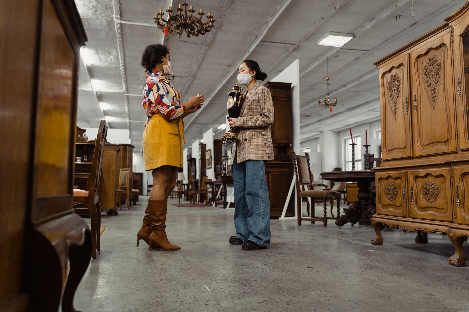 Two women wearing masks browsing antique furniture in a vintage store setting.