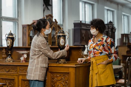 Two women wearing masks examine antique vases in a vintage store.