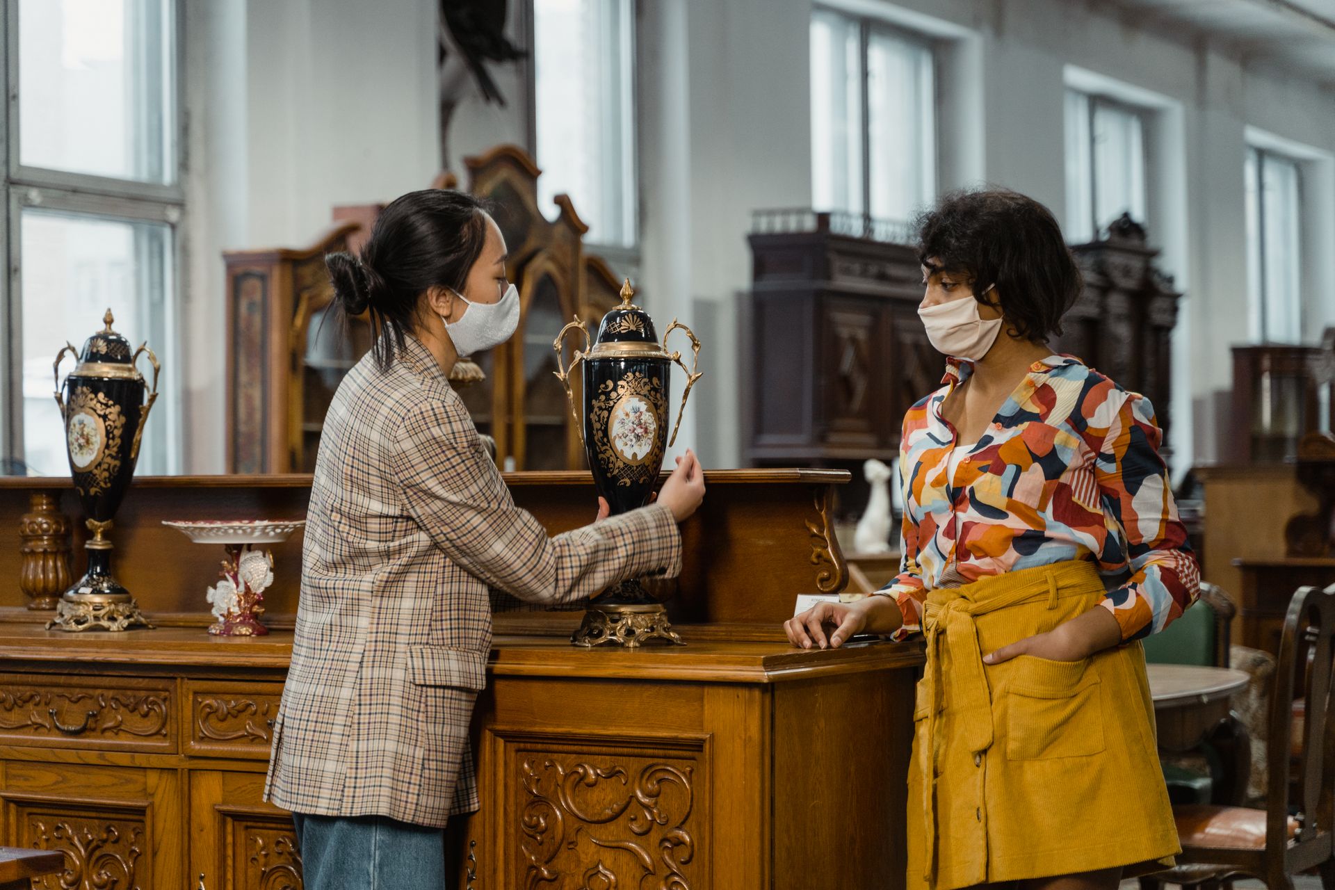 Two women wearing masks examine antique vases in a vintage store.