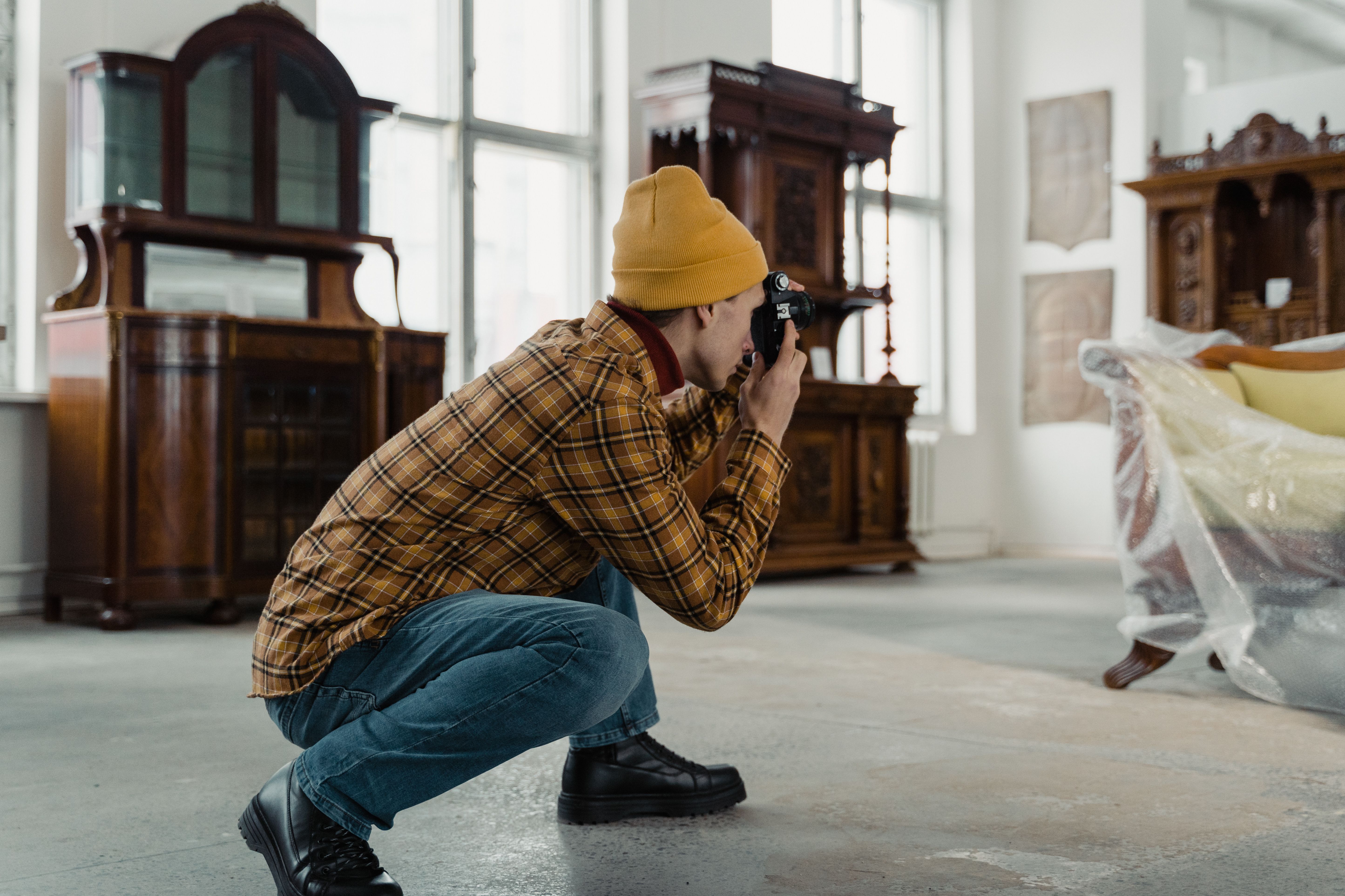 A person in a beanie photographs antique furniture inside a well-lit room.