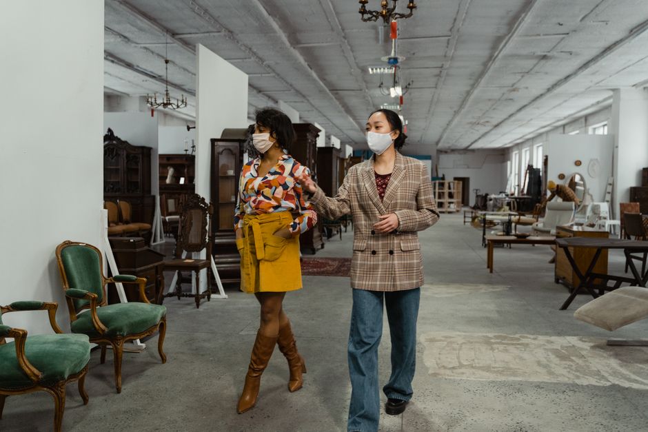 Two women wearing masks browse antique furniture in a spacious indoor store.