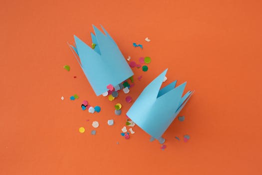 Two blue paper crowns and scattered colorful confetti on an orange backdrop, perfect for festive themes.