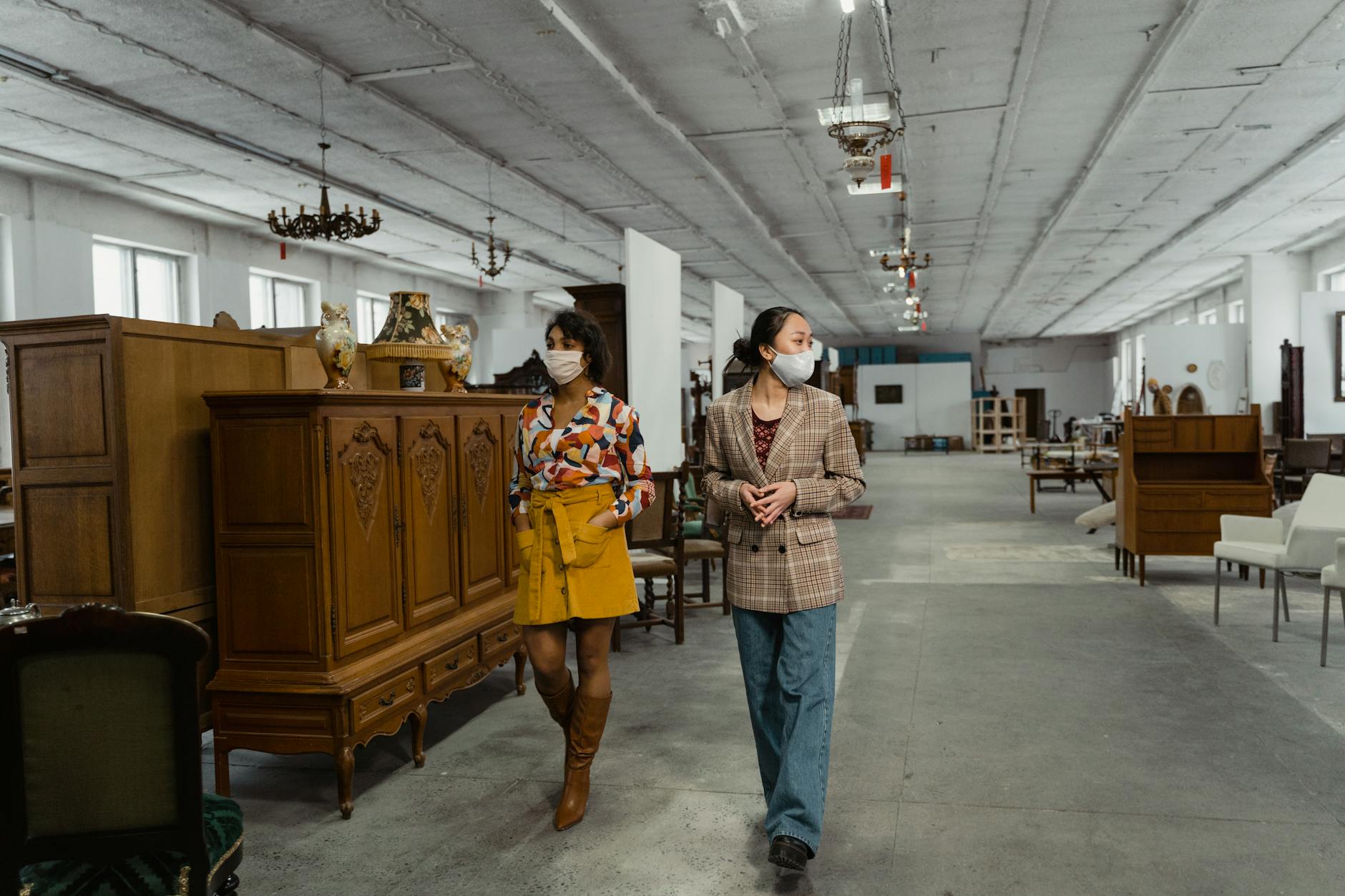 Two women in face masks explore a large store filled with vintage furniture and chandeliers.