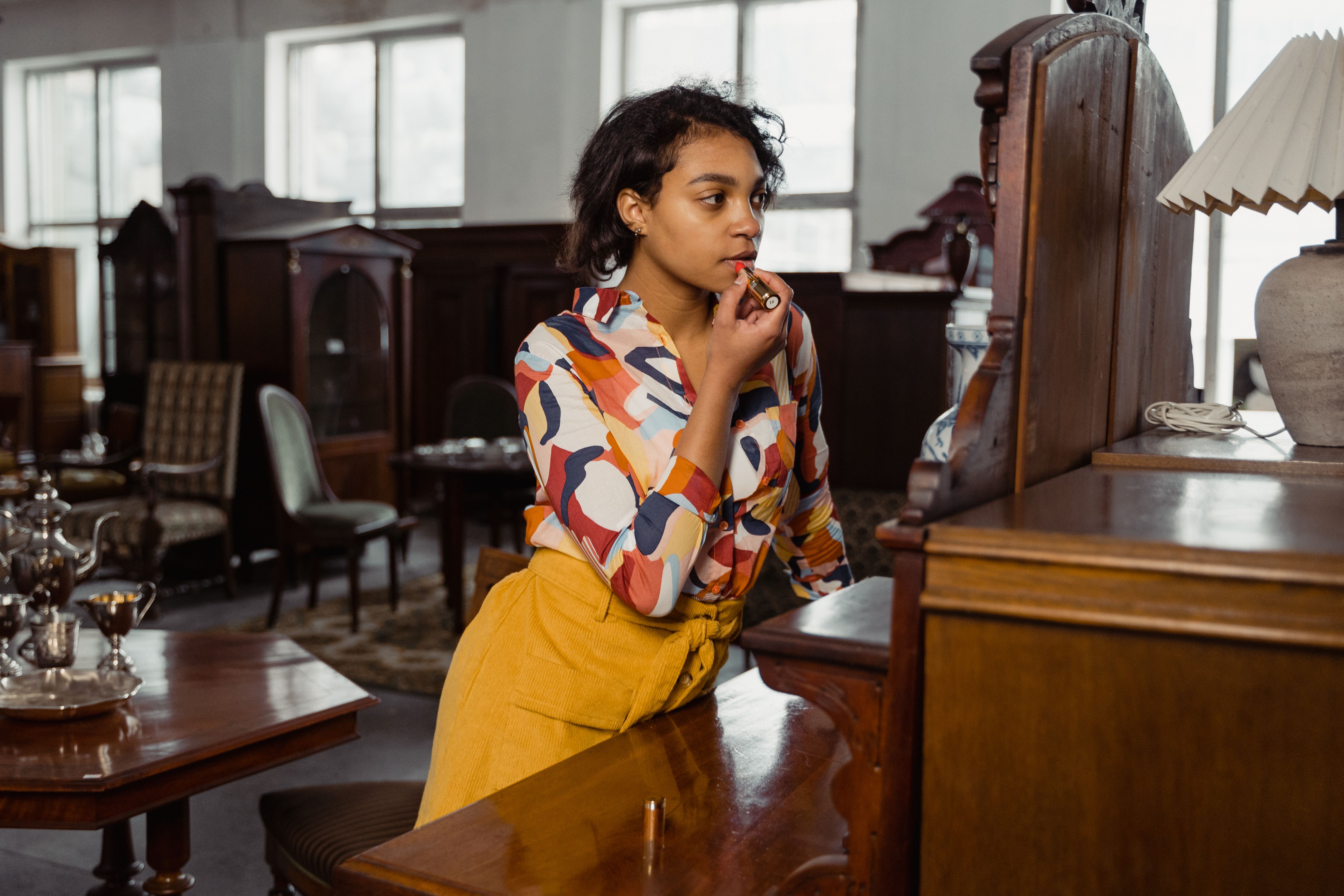Stylish woman applying lipstick while standing in a vintage furniture store with wooden furniture and decor.