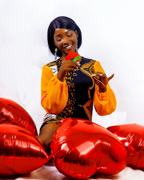 A Happy Woman Holding a Flower while Sitting beside Heart Shaped Balloons