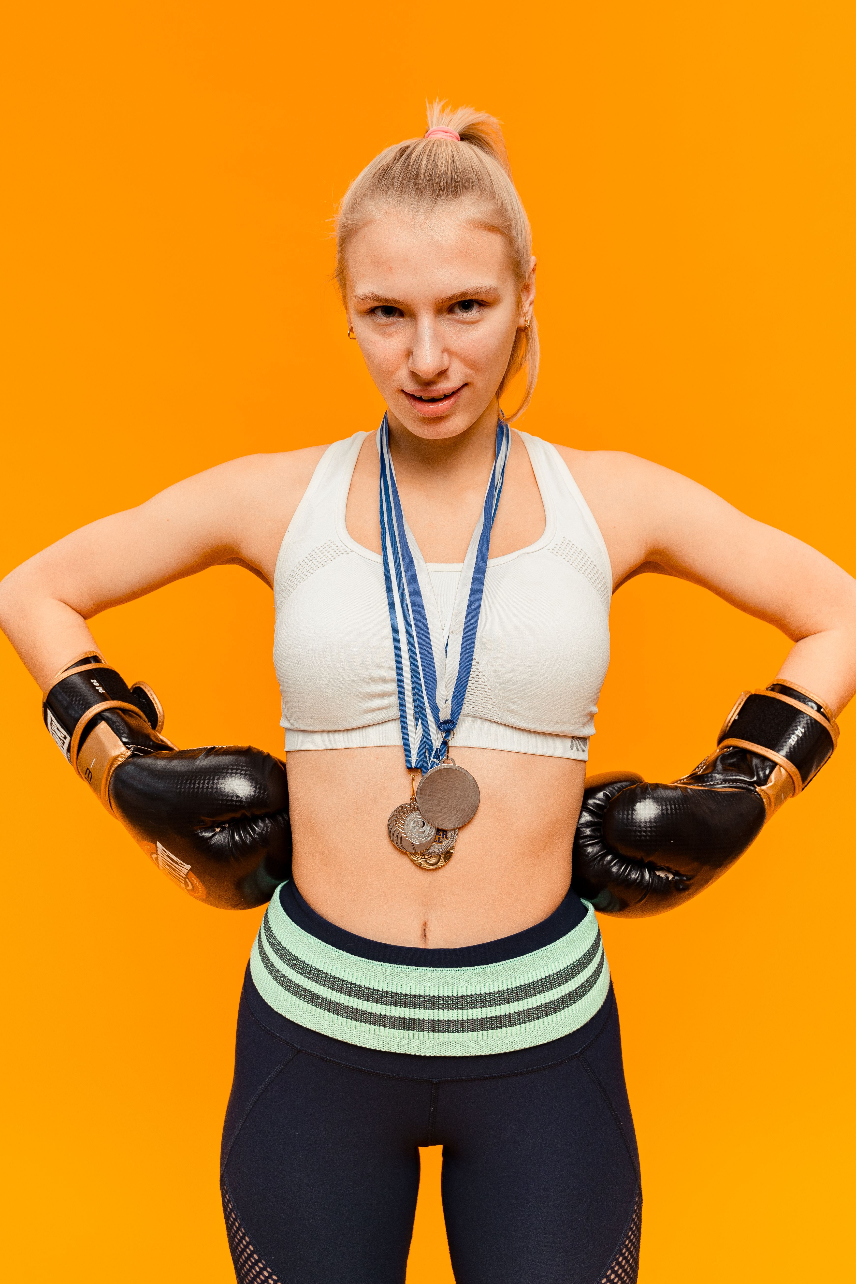 Confident female boxer poses with medals and boxing gloves against a vibrant orange backdrop.