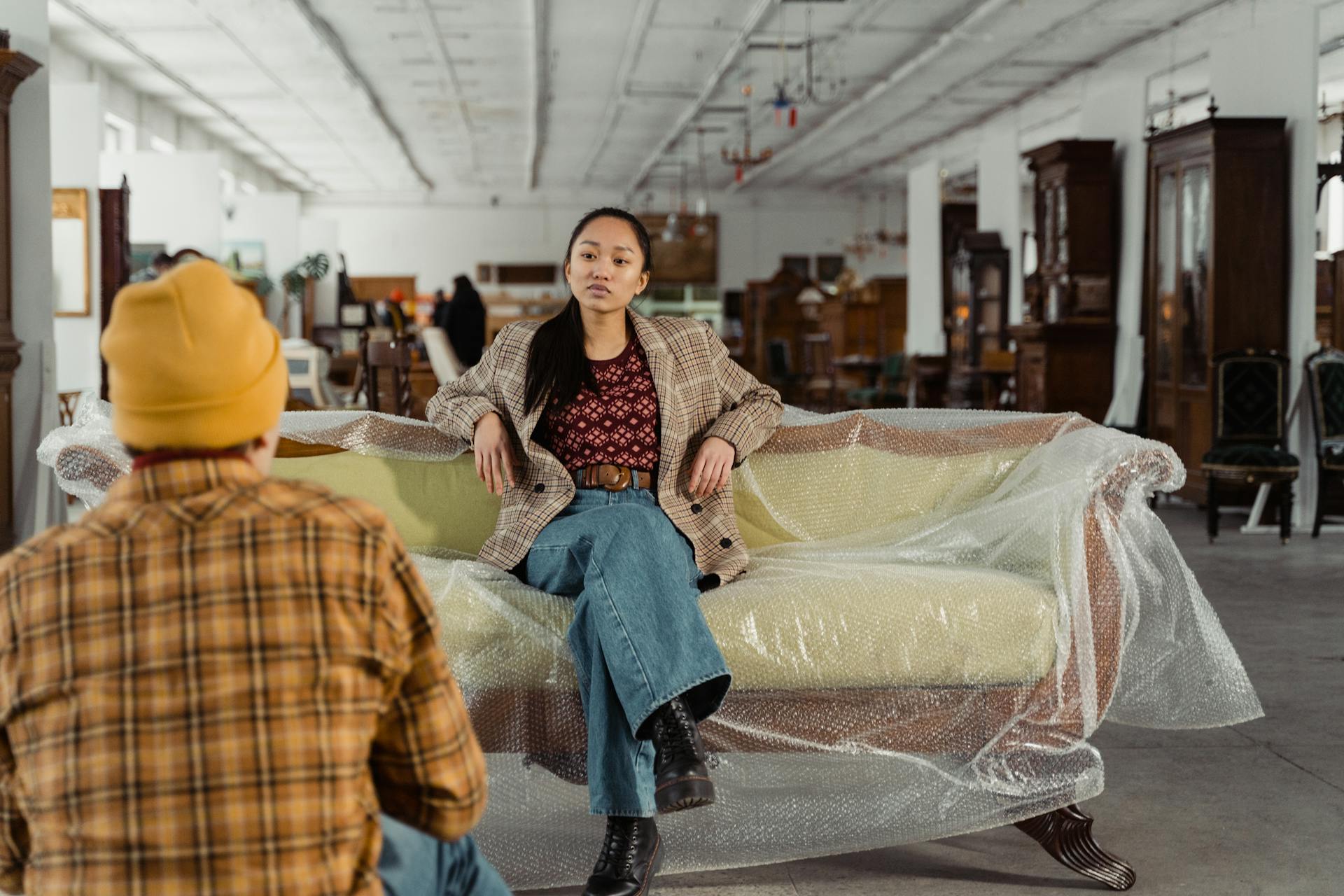 A woman in fashionable attire posing on a plastic-covered sofa in a vintage furniture shop.