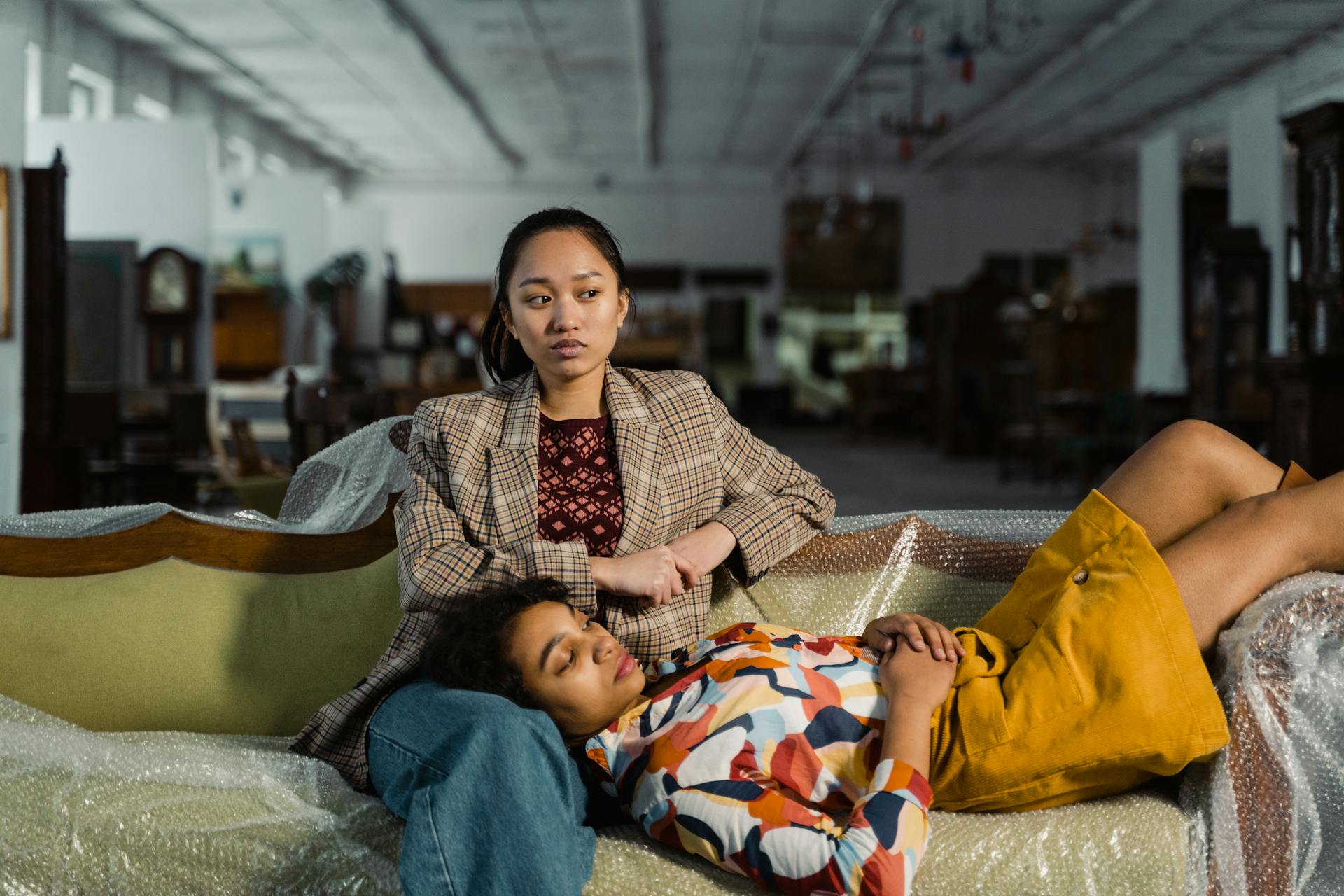 Stylish women posing in a vintage furniture store with trendy clothing and relaxed expressions.