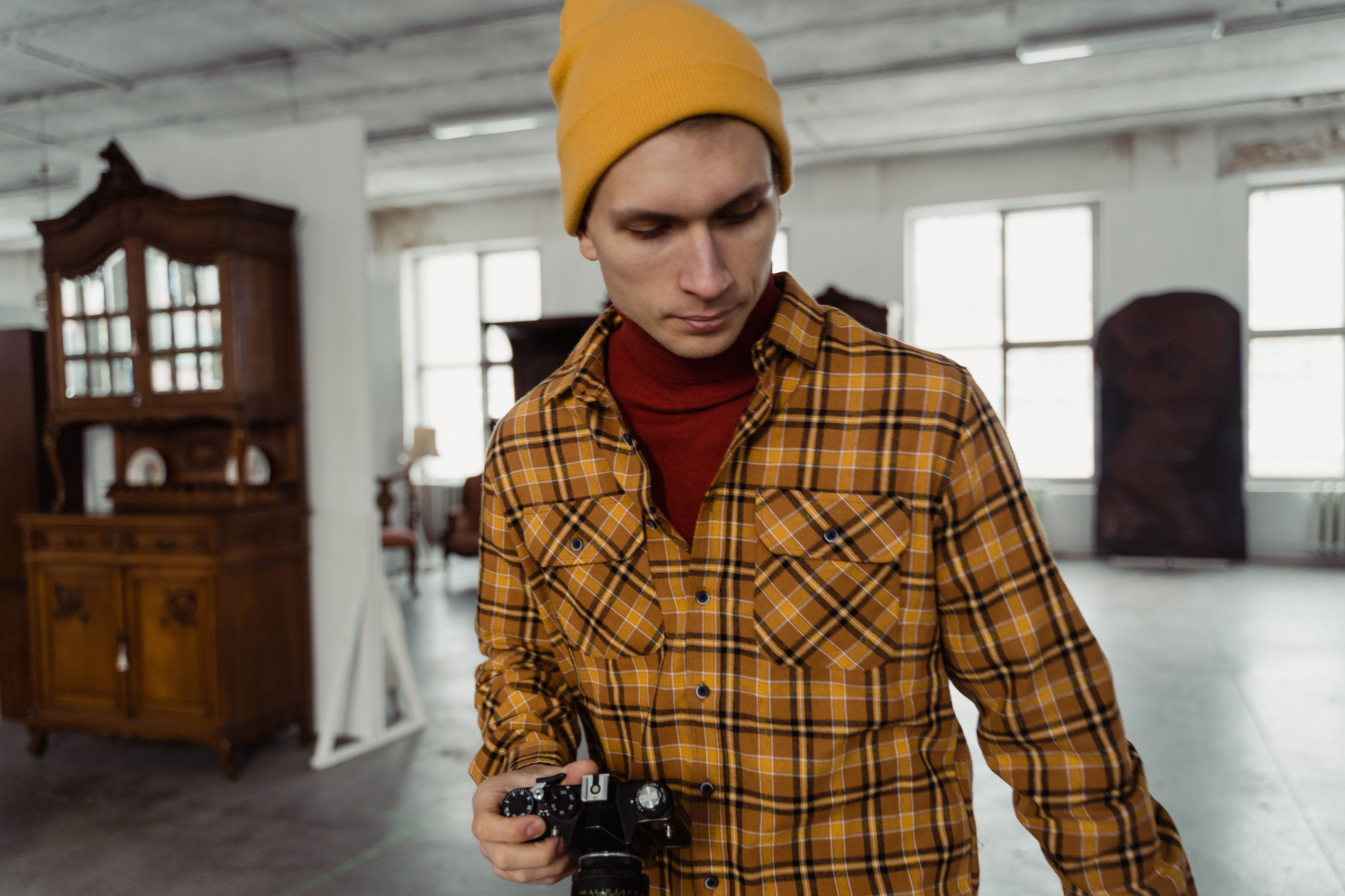 Photographer with vintage camera in an antique-filled shop wearing a yellow beanie and plaid shirt.