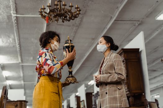 Two women in masks evaluating an antique vase in a shop, showcasing vintage fashion.