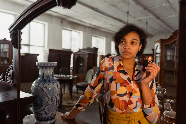 Woman In Colorful Shirt Holding Red Lipstick And Looking At The Vintage Mirror Beside Antique Vase