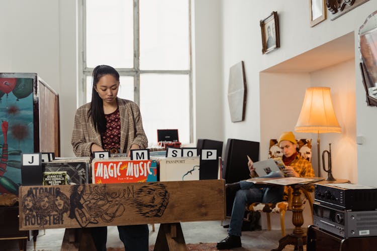 Woman Choosing Vinyl Record In Vintage Store Beside Man Reading A Magazine While Sitting In Armchair