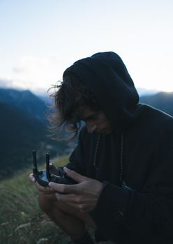 A young man in a hoodie operates a drone controller amidst mountains during the day.