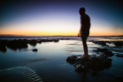 Silhouette of Man Standing on Rock Surrounded by Body of Water