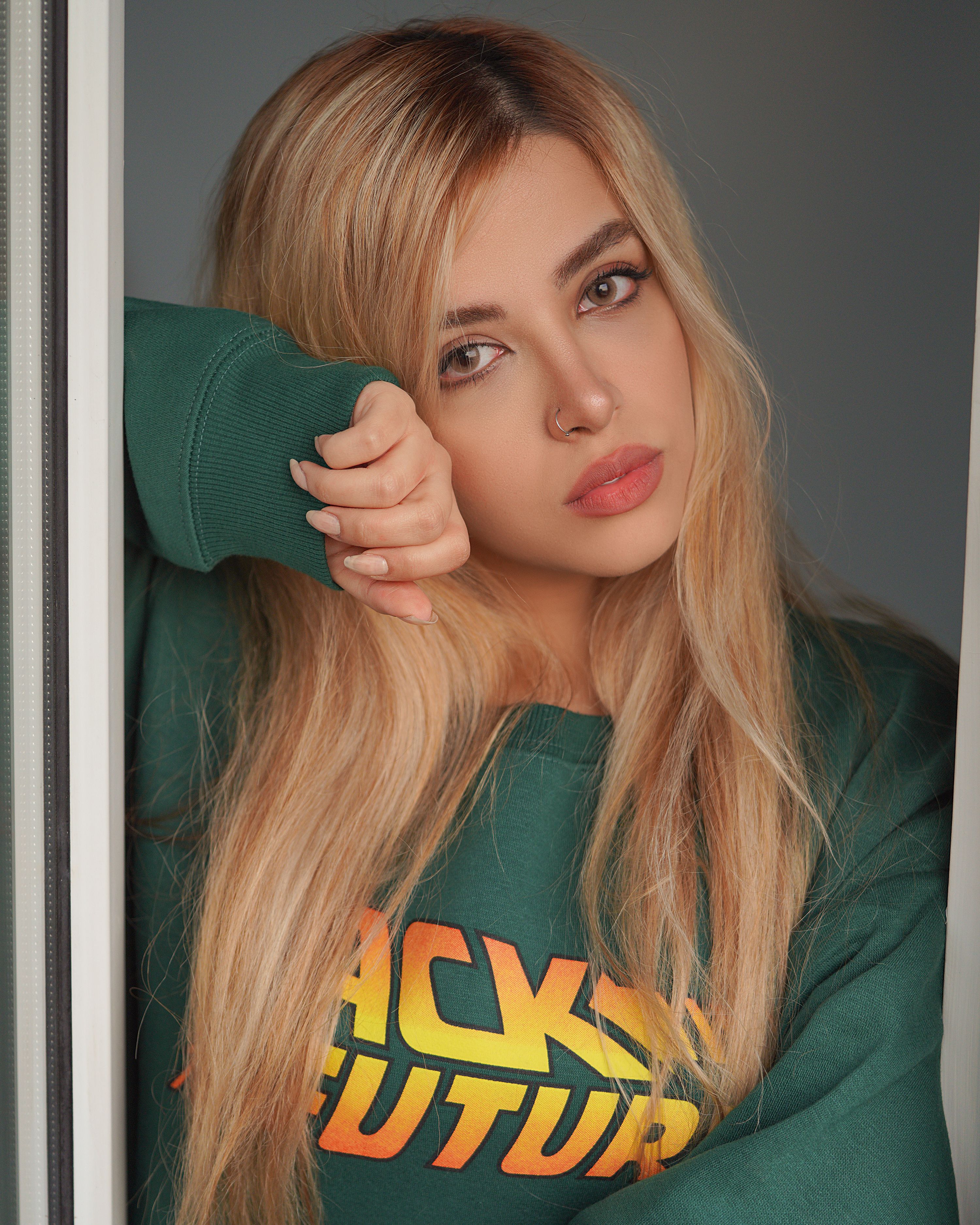 Close-up portrait of a blonde woman with a nose piercing in a green sweater, looking thoughtfully.