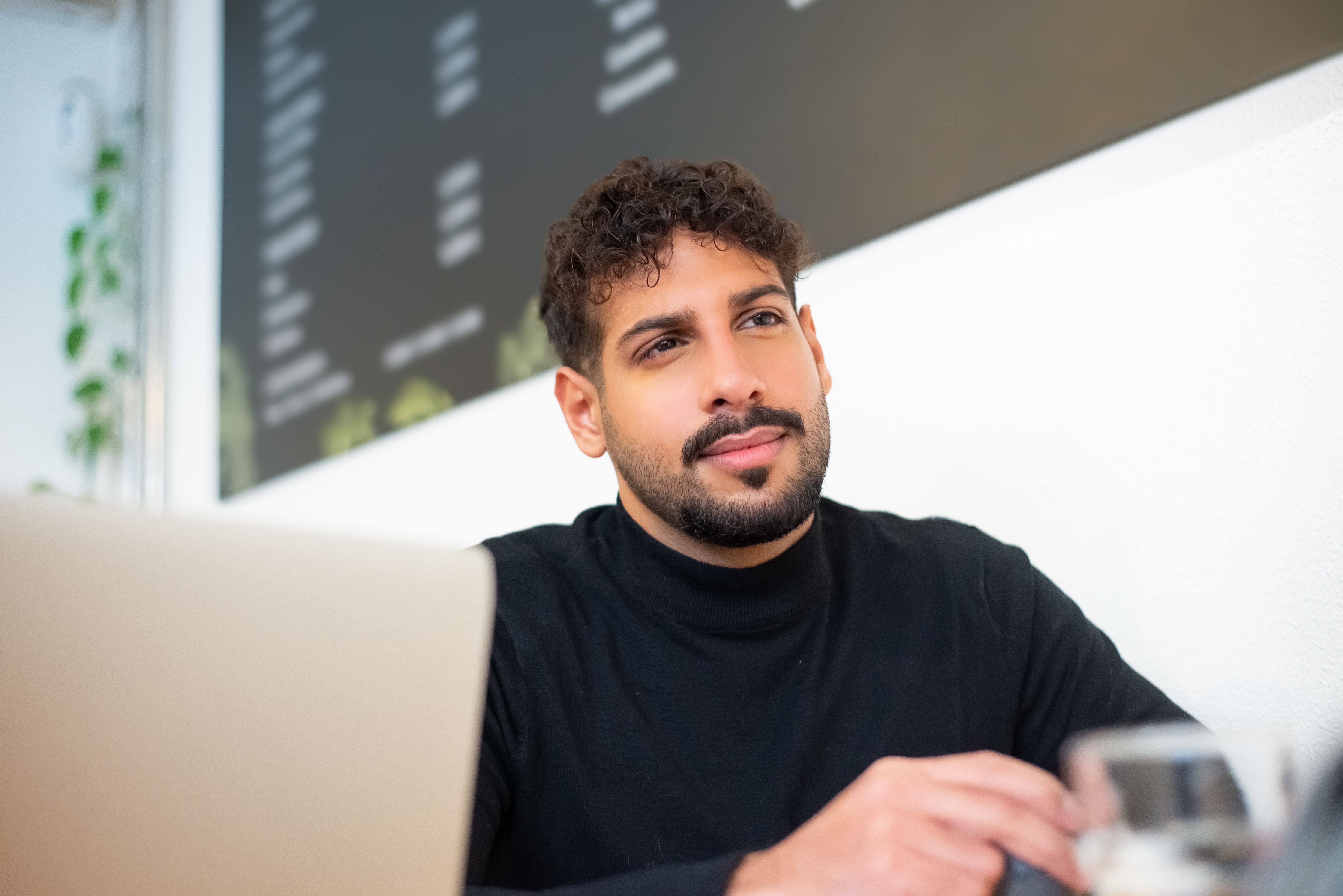Bearded man in black sweater sitting at a cafe table with a laptop, thoughtful expression.