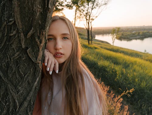 Young female standing behind tree trunk and looking at camera while spending sunny day in riverside