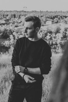 Black and white of stylish male with folded arms wearing black outfit and looking away thoughtfully while standing in countryside