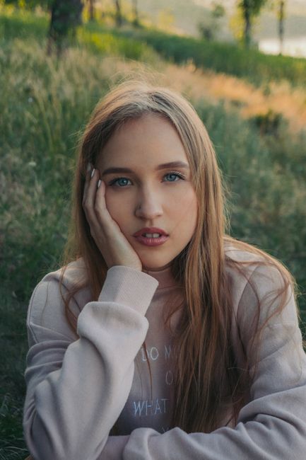 Portrait of young blonde female holding hand on cheek and looking at camera while spending summer evening in countryside