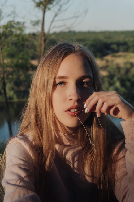 Portrait of calm pensive female with long straight hair looking at camera while resting on sunny meadow near river