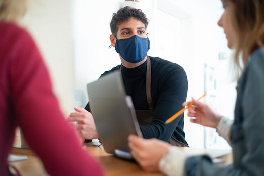 Coworkers in a business meeting wearing facemasks, discussing a project in a modern office setting.