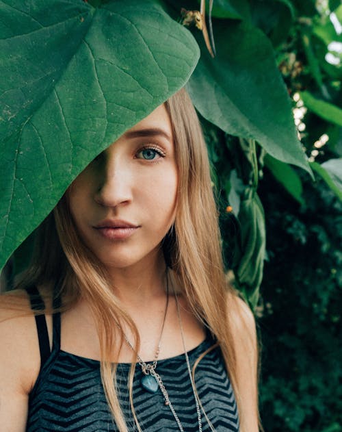 Portrait of charming blond lady in stylish outfit with blue eyes looking at camera while hiding under green foliage