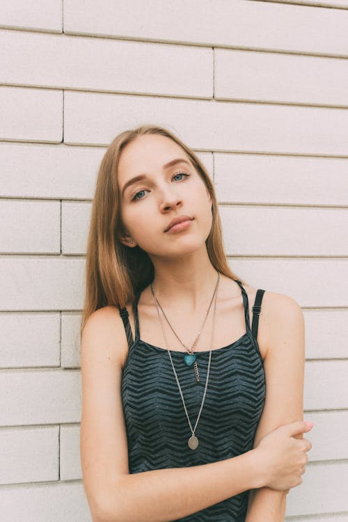 Young confident slender female touching arm and looking at camera near modern brick white wall