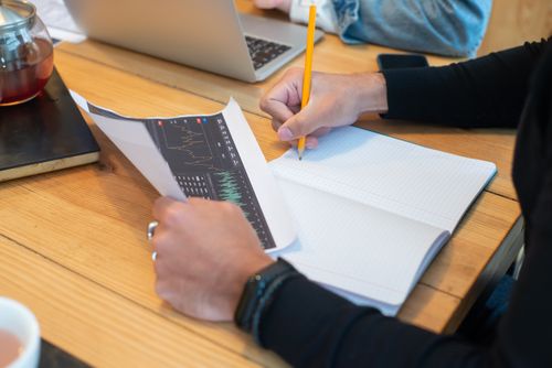 Free Close-up of hands writing notes on notebook during business meeting with laptop and charts. Stock Photo