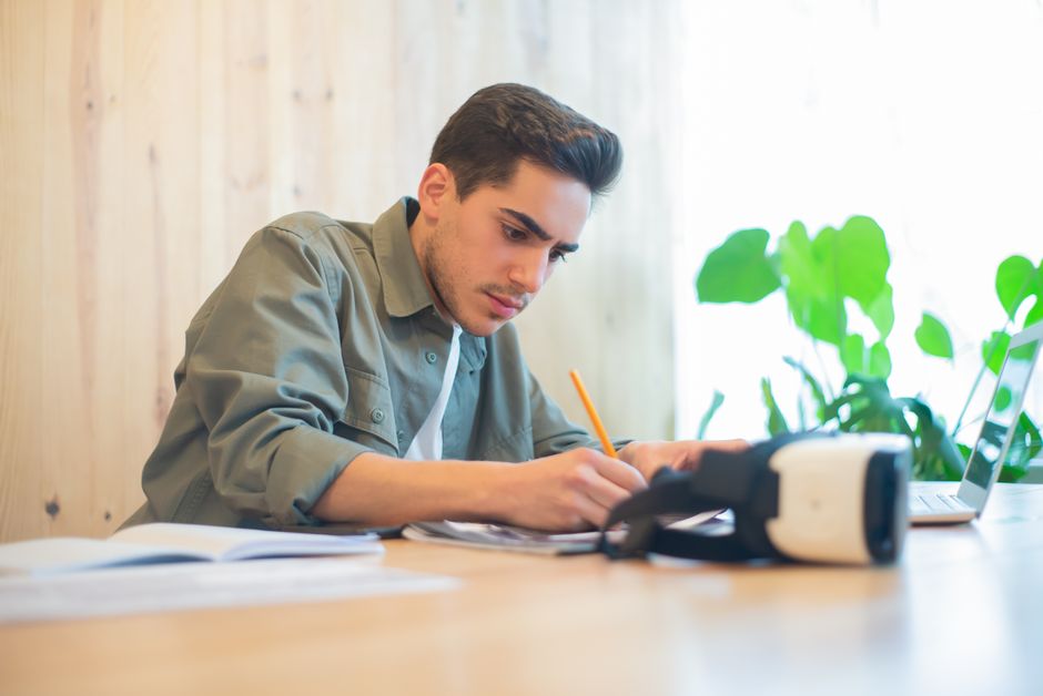 Focused young man working at a desk with VR goggles, laptop, and papers in a sunlit office. Focused young man working at a desk with VR goggles, laptop, and papers in a sunlit office.
