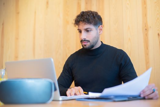 Focused businessman with beard working on a laptop, reviewing documents in a modern workspace.