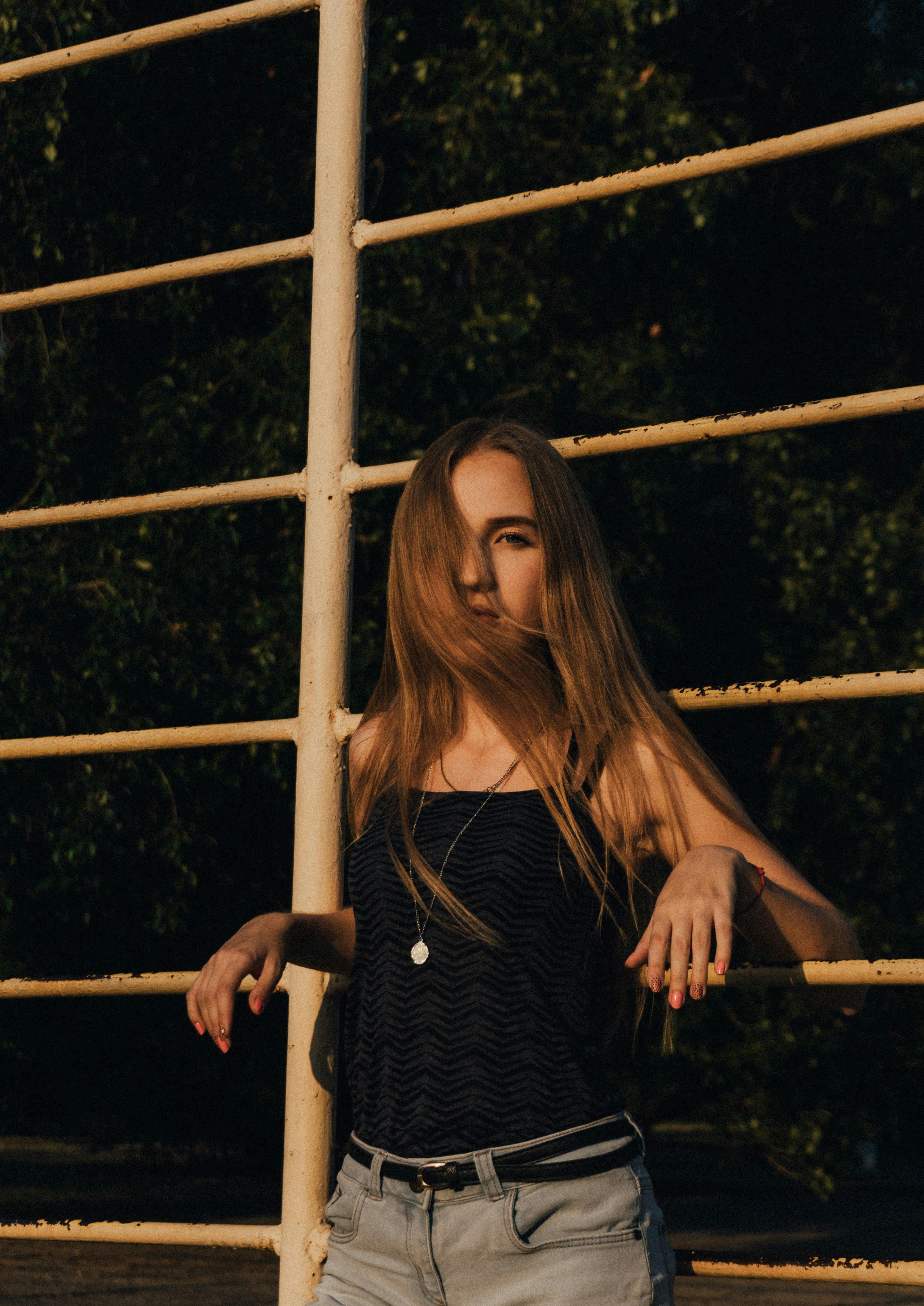 Young female with long fair hair in stylish clothes looking at camera while leaning on iron railing