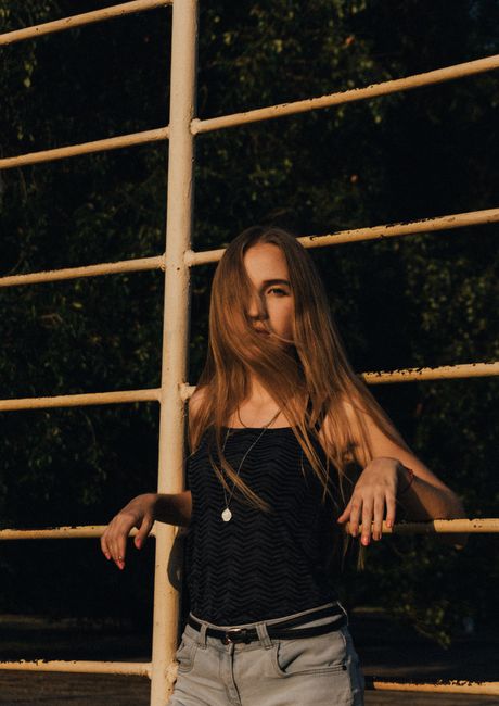 Young female with long fair hair in stylish clothes looking at camera while leaning on iron railing