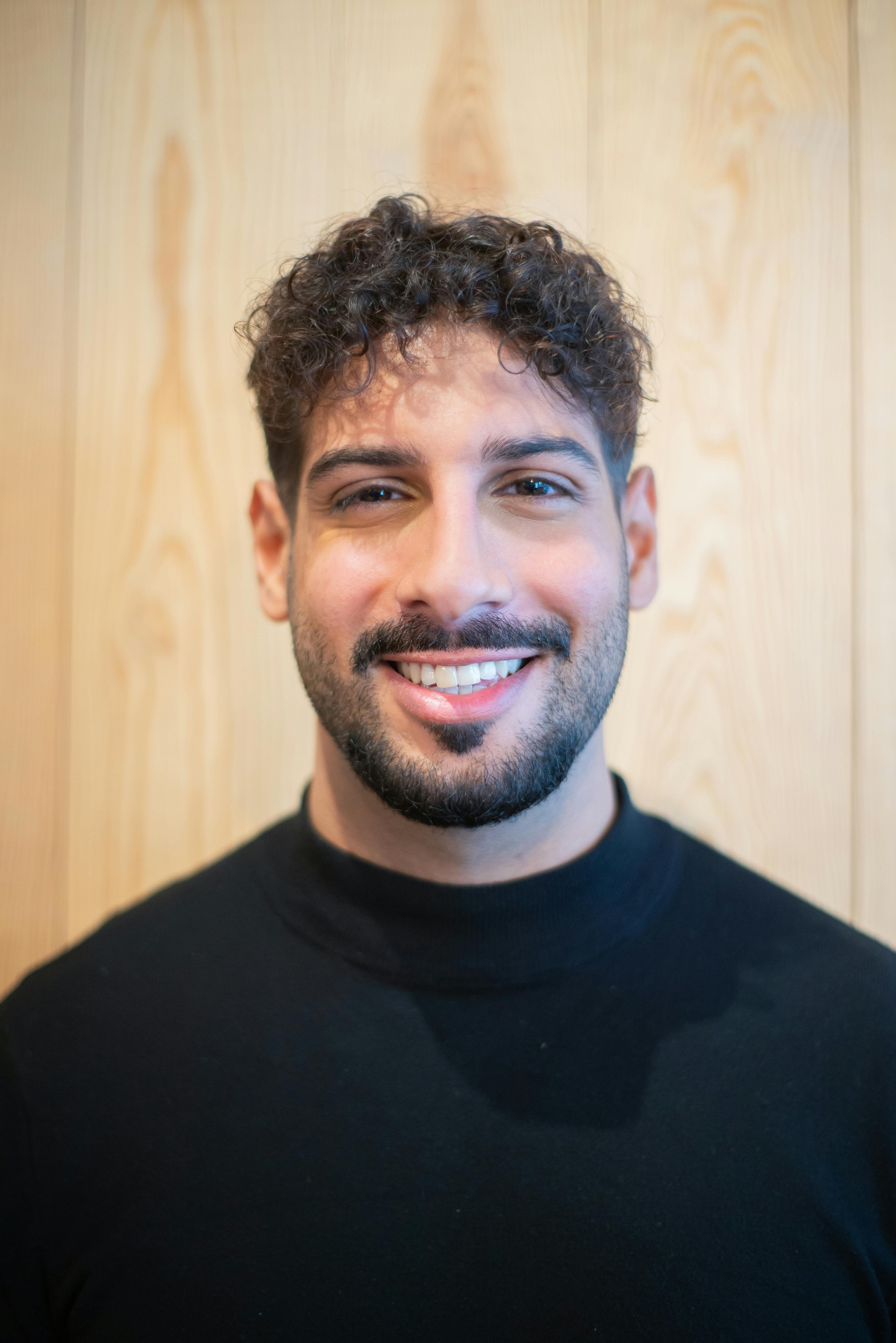 Close-up of a smiling man with curly hair against a wooden background.