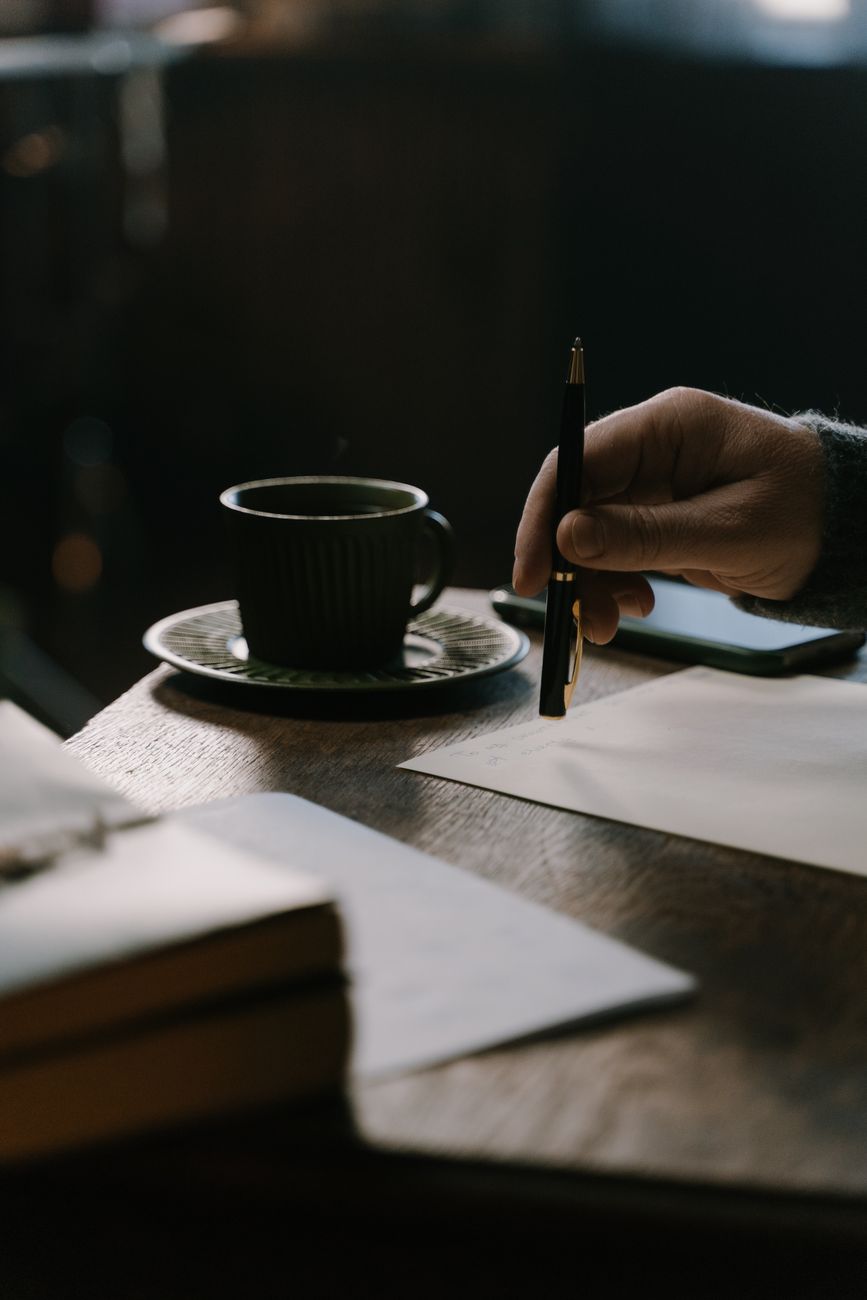 Hand holding a pen over a journal beside coffee on a wooden desk
