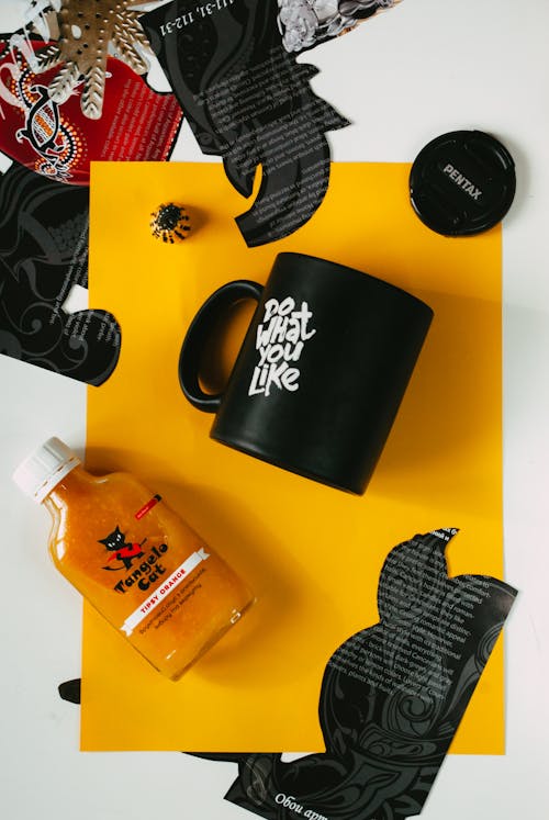 Top view of bottle of orange sauce and black ceramic mug with Do What You Like inscription arranged on white table with various magazine clippings