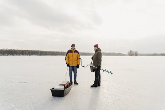 Two adults standing on a frozen lake with ice fishing gear during winter.