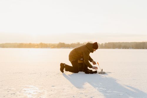 Free A man kneeling on a frozen lake while ice fishing during a sunny winter day. Stock Photo