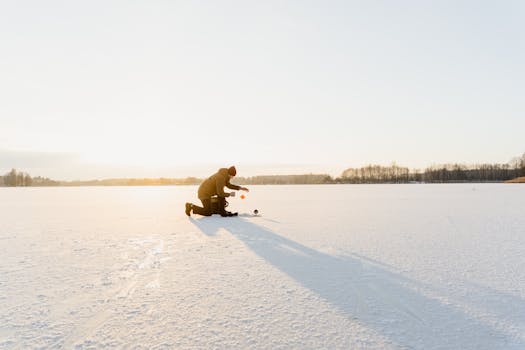 A lone fisherman ice fishing on a frozen lake during winter sunrise.