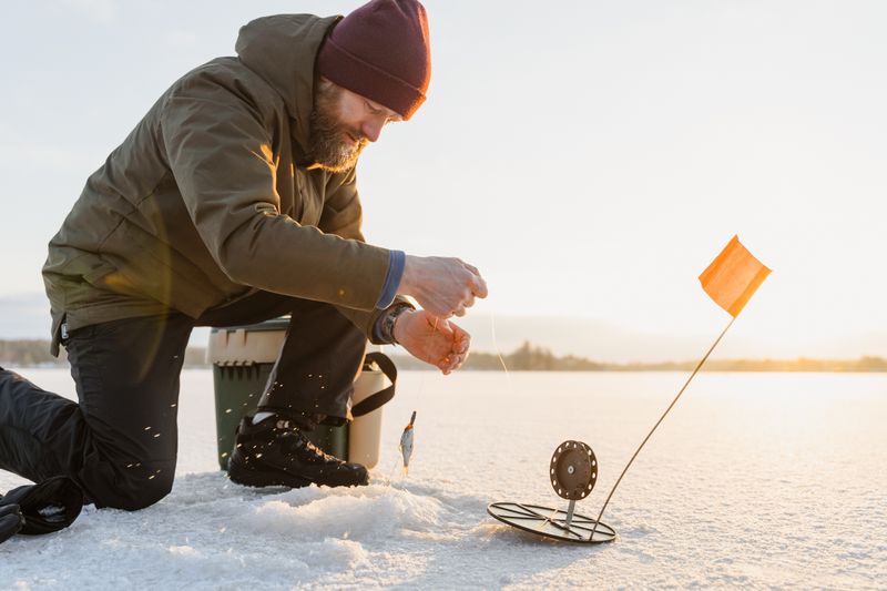 Angler ice fishing at sunrise on a frozen lake, rod in hand, line dropping through the ice hole